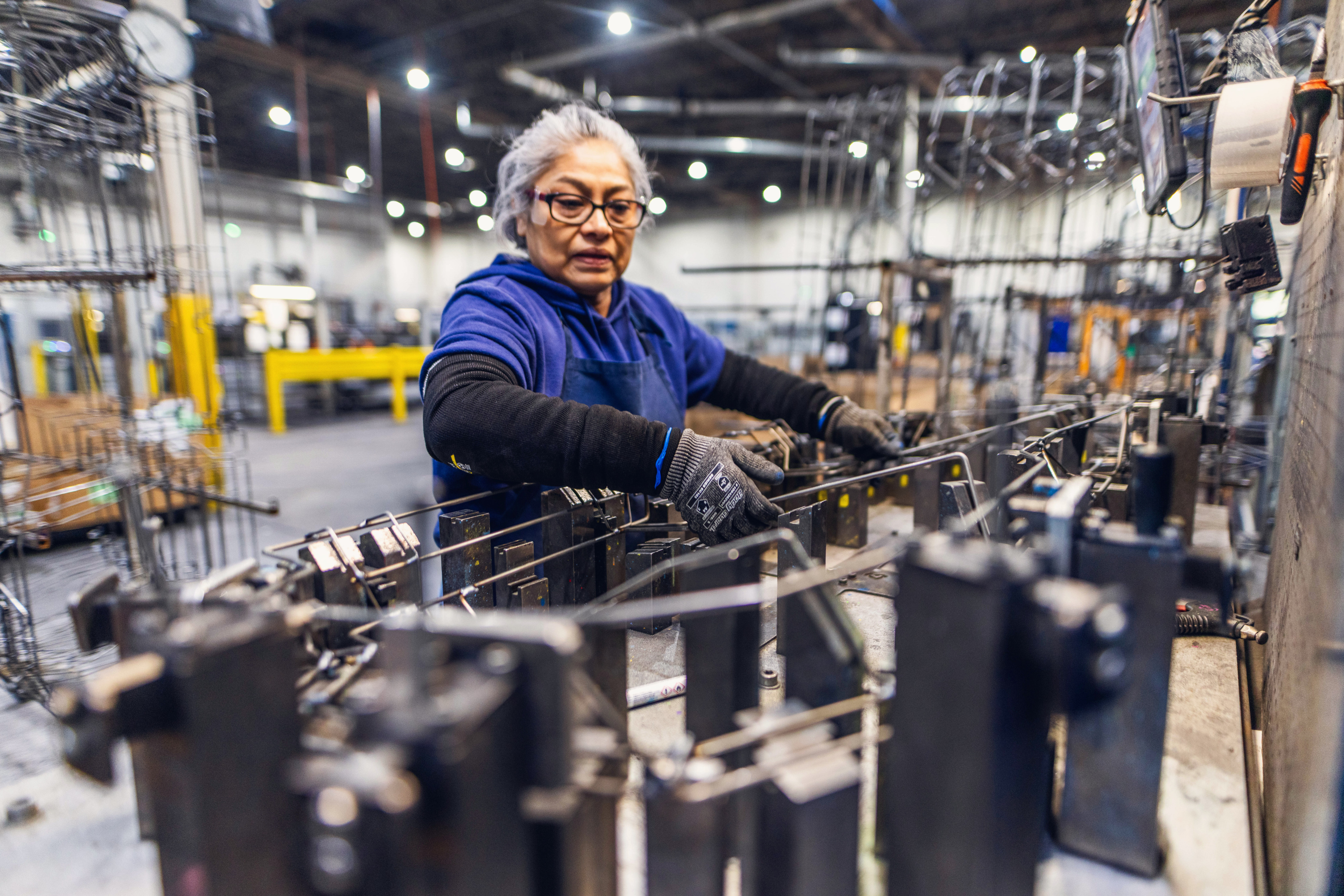 Older woman in safety glasses and blue hoodie operating a conveyor line in a busy factory.